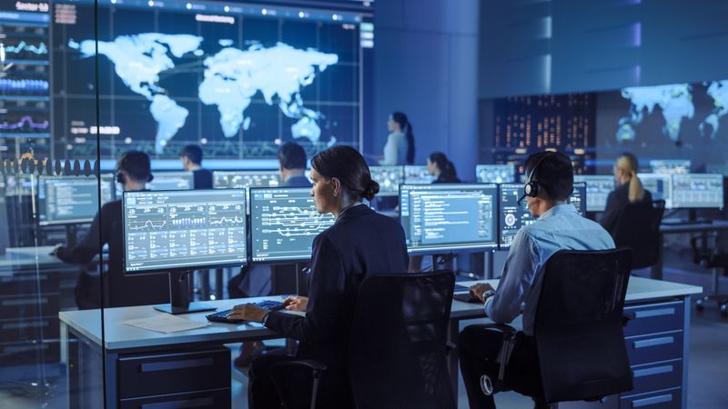 Diverse Team Monitors Global Data in High-tech Operations Center. Multiple Screens Display Charts and World Maps.
