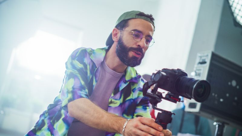 Focused Young Male Cinematographer With Beard, Adjusting Settings On A Modern Camera During An Indoor Film Shoot, Wearing A Colorful Jacket And Glasses. Creativity And Professionalism In Filmmaking.