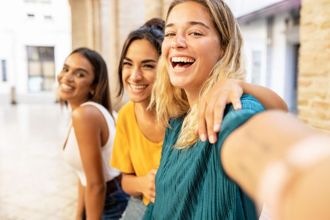 Portrait of three cheerful multi-ethnic female friends taking selfie with phone on city street