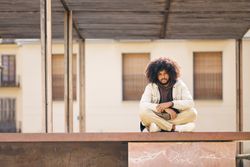 Young man with afro hair sitting cross-legged on a step on a quiet city street looking at the camera