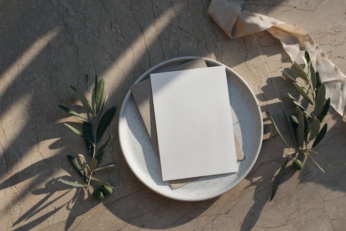 Blank greeting card, invitation mock up on plate surrounded by olive tree branches in long shadows