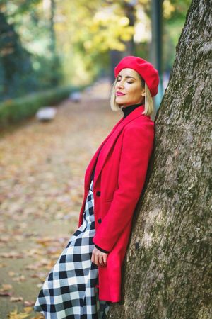 Side view of dreamy female with closed eyes wearing elegant red outerwear leaning on tree trunk in autumn park