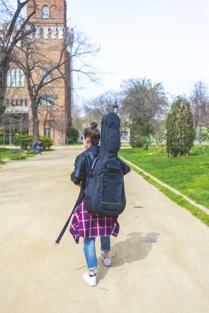 Rear view of a young hipster woman walking in a park in sunny day while carrying a guitar on back