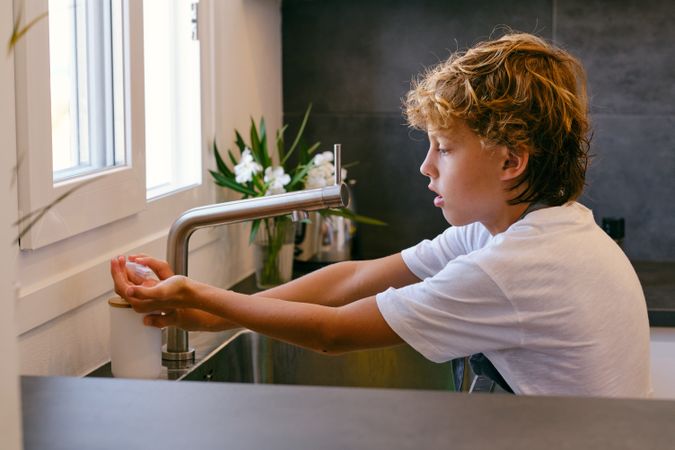Boy applying liquid soap on hands above sink in kitchen
