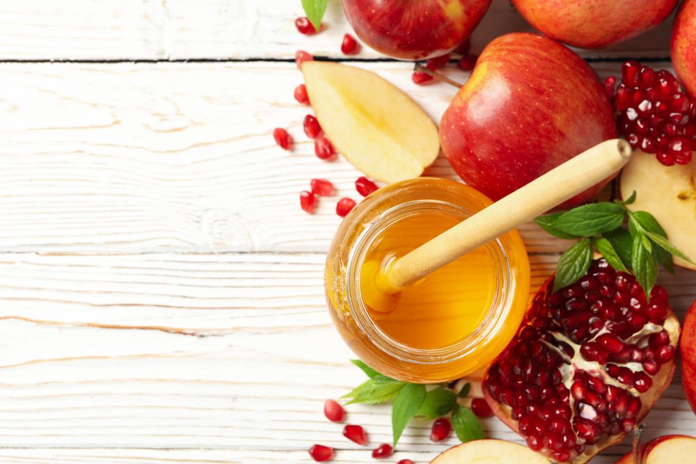 Top view of glass honey pot with dipper, pomegranate seeds, and an apple on side with copy space