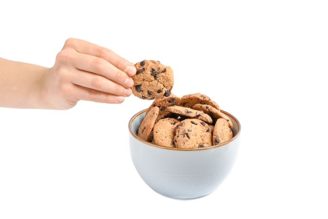 Young woman holding tasty chocolate chip cookie over bowl on blank background, closeup