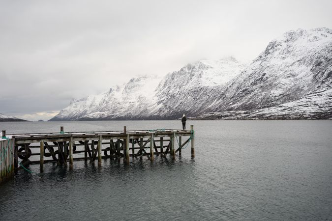 Lonely tourist standing on wooden pier in middle of calm lake in winter