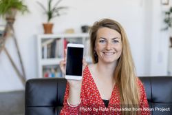 Happy woman showing a blank smart phone screen sitting on a sofa at home 0Pjzeg