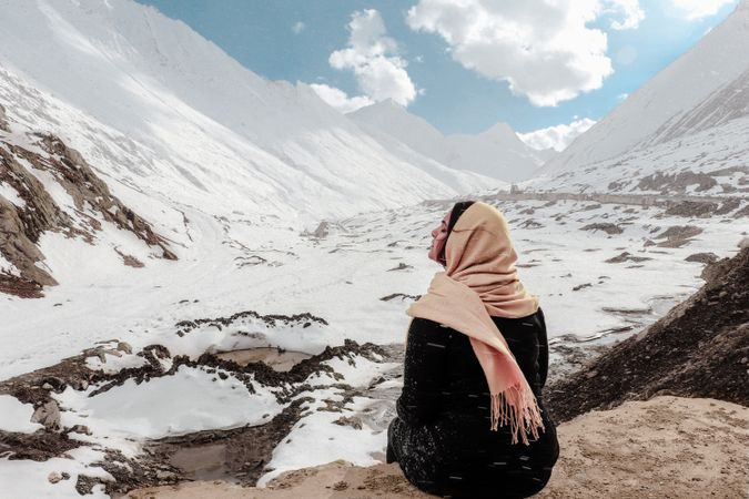Back view of woman with pink headscarf sitting on a rock looking at snow-covered mountains