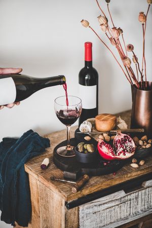 Rustic setting of hand pouring wine, cheese, long candles, with pomegranate, and dried poppies