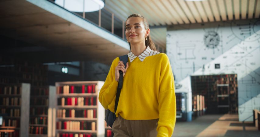 Female Student Walking in an Empty Public Library with Bookshelves. Young Smart Woman Carrying a Backpack with Textbooks to Prepare and Study for Exams. Low Angle Portrait