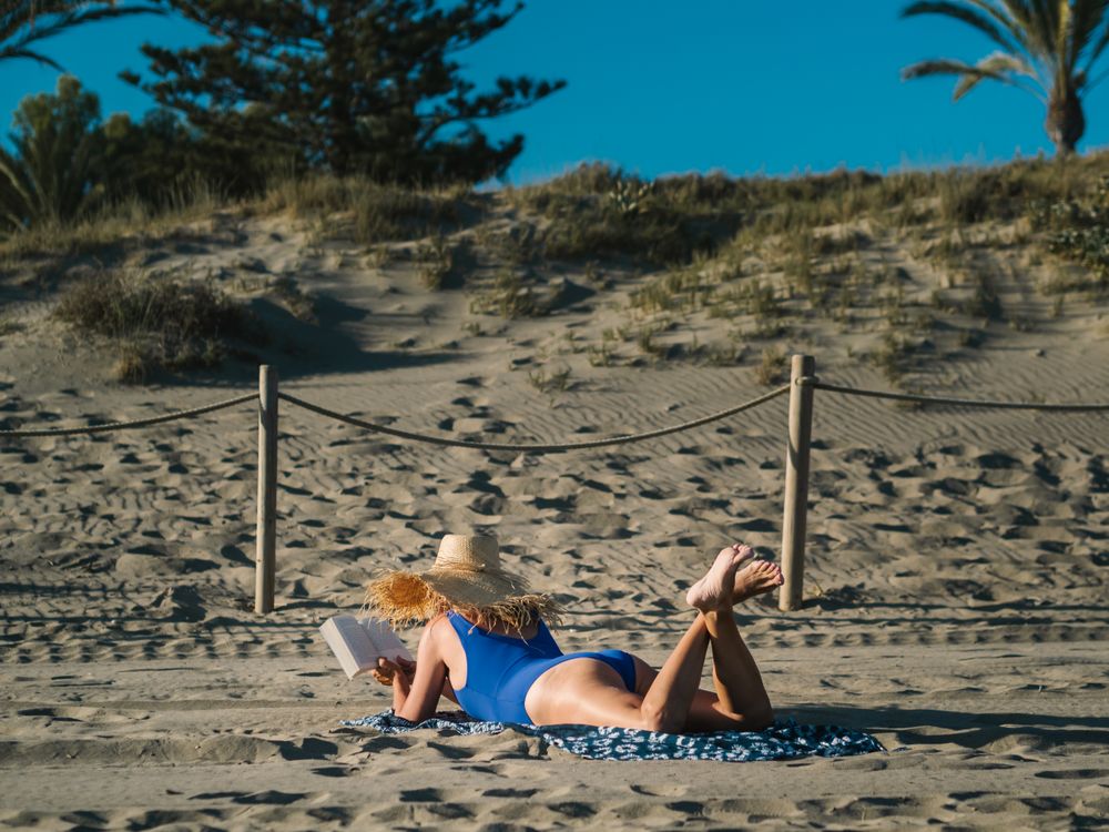 Sandy beach and woman in big hat lying and reading book