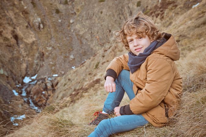Smiling boy in warm clothes sitting on grassy hill