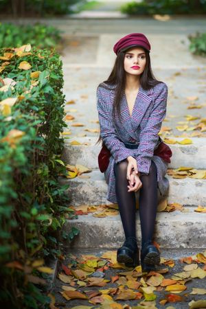 Female in warm winter clothes sitting among scattered fall leaves on park steps looking to her side