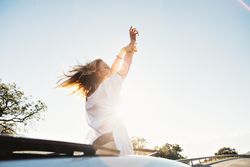 Rearview of woman waving out of sunroof on sunny day with her arms up