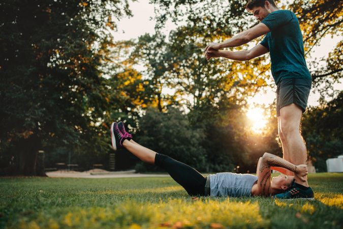 Woman lying on grass and stretching with support from her personal trainer