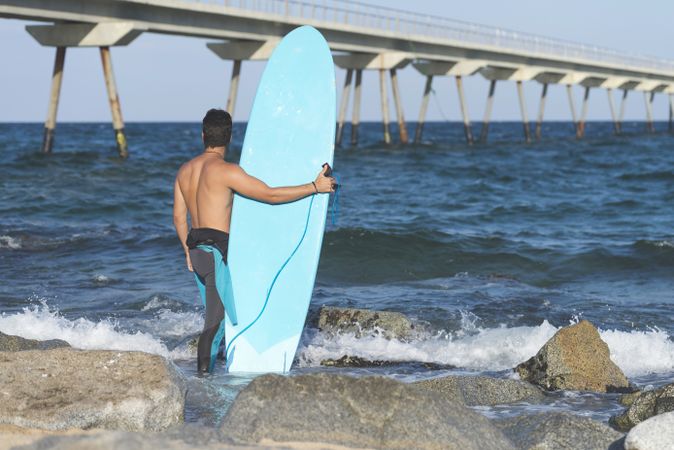 Male surfer with blue board standing in the ocean around a rocky beach