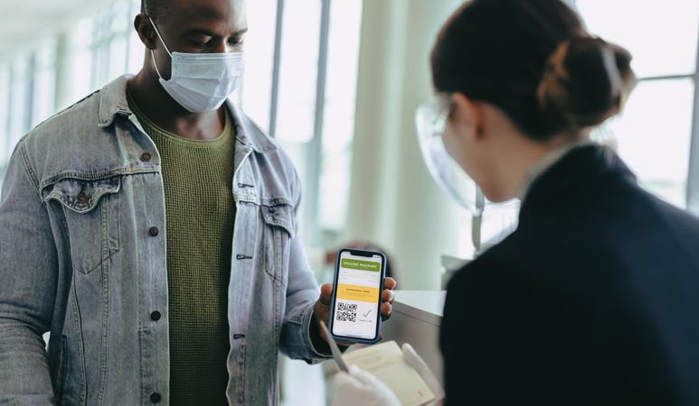 Man with face mask holding mobile phone to show vaccine passport to airport staff