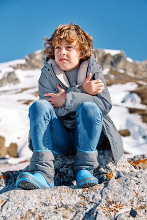 Blond boy sitting on snowy rocky hill