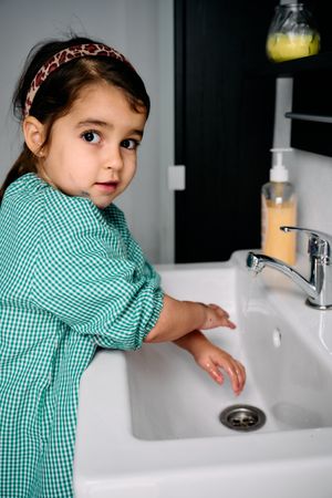 A little brunette girl with a green baby is washing her hands in the bathroom at home.