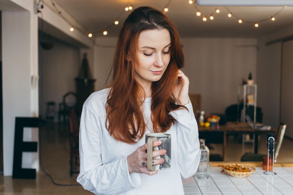 Woman plays with hair while holding a mug of a warm drink in her apartment