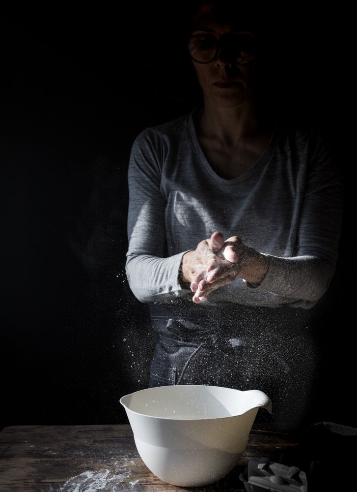 Woman clapping hands in flour near bowl and pack of eggs on table