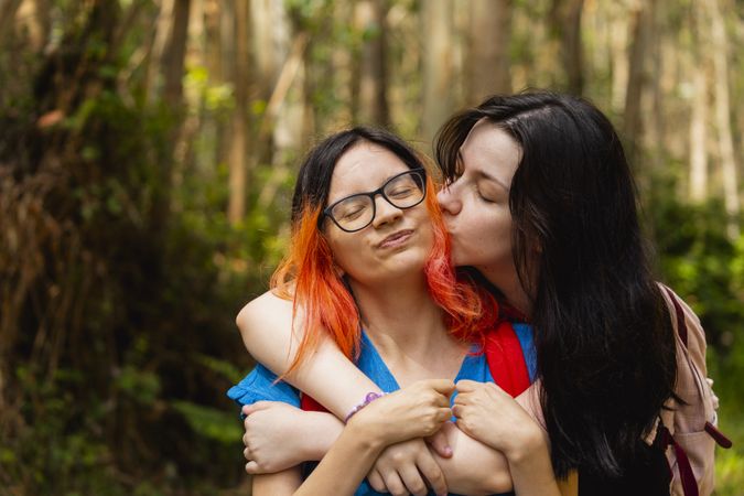 A young woman gives her friend a kiss while hugging her, during a walk in the woods