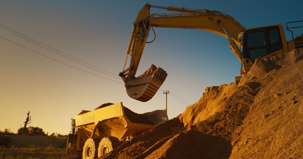 Construction Site On Sunny Evening: Industrial Excavator Loading Sand Into A Truck. The Process Of Building New Apartment Block. Workers Operating Heavy Machinery To Complete New Real Estate Project.