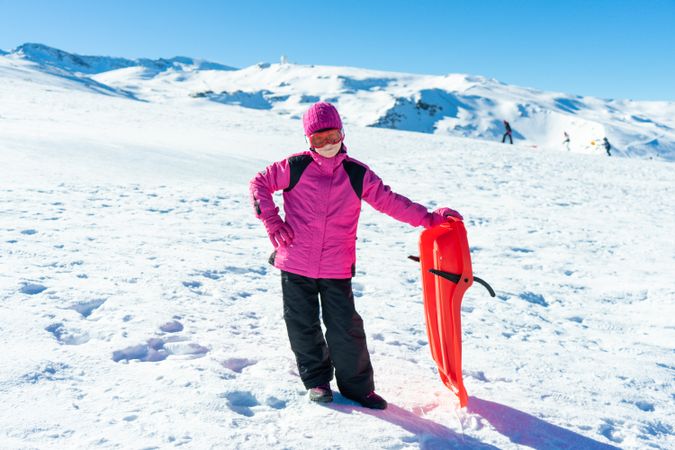 Child in pink snow suit standing on hill with sled at resort