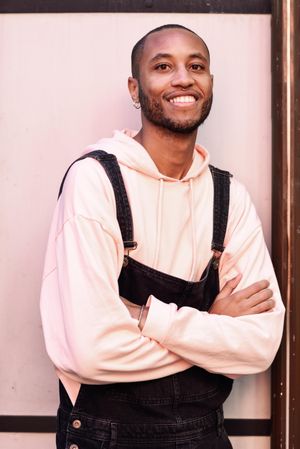Smiling male in denim overalls and earrings with arms crossed against a pink wall