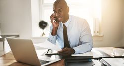 Man in suit and tie smiling on phone while working on his laptop with documents in hand
