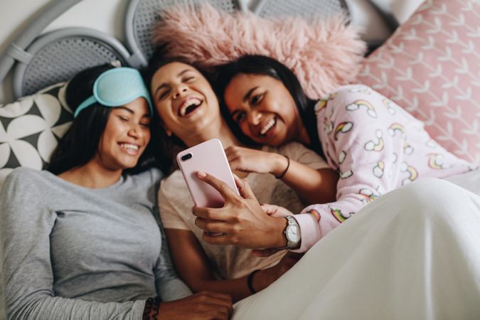 Girls taking selfie using mobile phone lying on bed with friends at sleepover