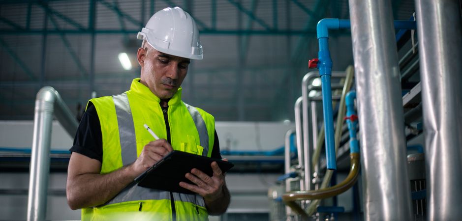 A male industrial engineer wearing a safety helmet and reflective vest is inspecting equipment in a factory. He is using a digital tablet to review data. Background show industrial pipes and machinery