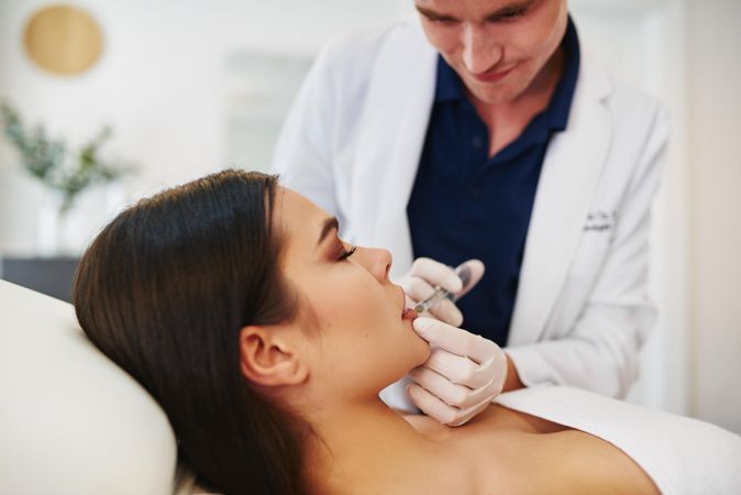 Side view of male dermatologist injecting treatment into brunette patient’s lips