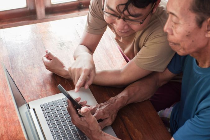 Happy adult son teaching older father using laptop and smartphone