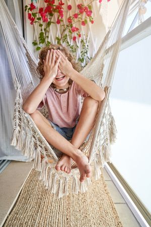 Cheerful boy sitting in hanging chair on balcony with blooming flowers