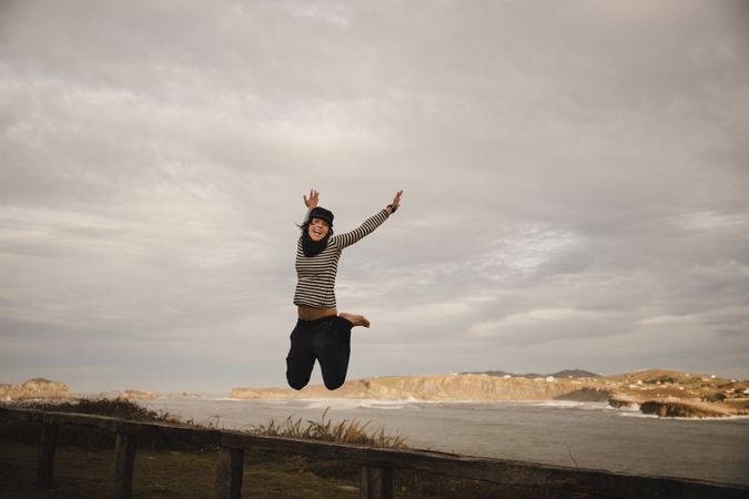 Young woman in hats having fun on seat on coast near waving sea and cloudy sky