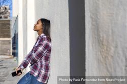 Female standing in the sun while leaning on wall listening to music on smartphone 0yZMG5