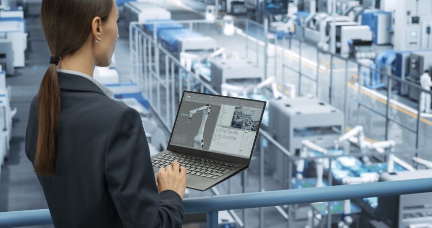 Young Female Engineer Standing on a Platform, Using Laptop Computer at an Electronics Factory. Machines are Undergoing Maintenance, Technician Monitoring Updates Through Online Software
