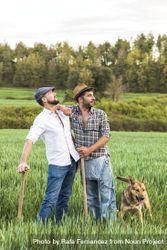 Two men in plaid shirt standing in long grass with forest in background with dog 56Gl3z