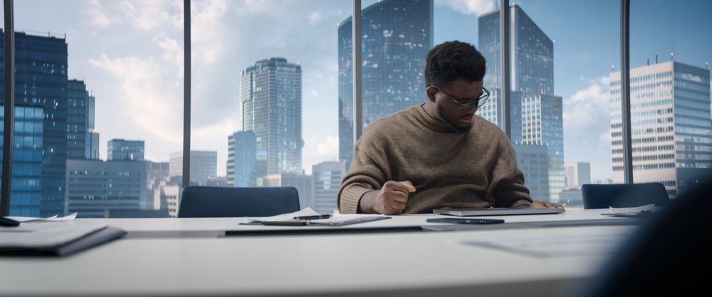 Confident Black Businessman Sitting at Desk in Office, Using Laptop Computer, Next to Window with Big City with Skyscrapers View.