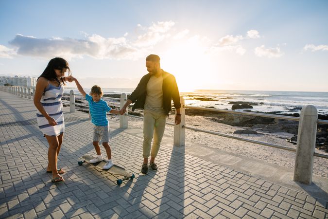 Couple helping their child learn skateboarding by the sea side holding his hands