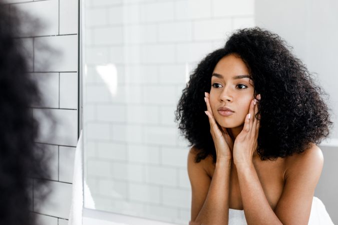 Black woman with curly hair touching her cheeks in the mirror