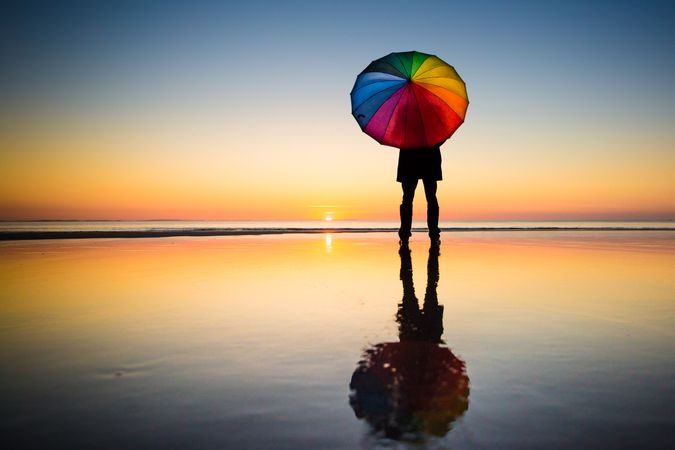 Silhouette of person holding  rainbow umbrella standing on seashore during sunset