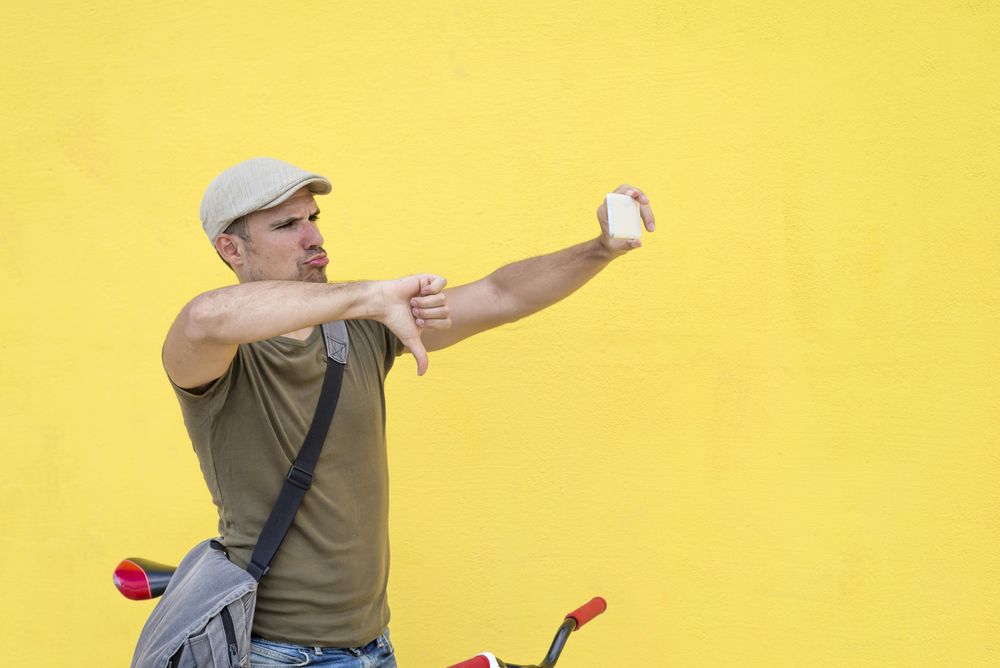 Male in hat and sunglasses taking selfie giving thumbs down on phone while sitting on bicycle