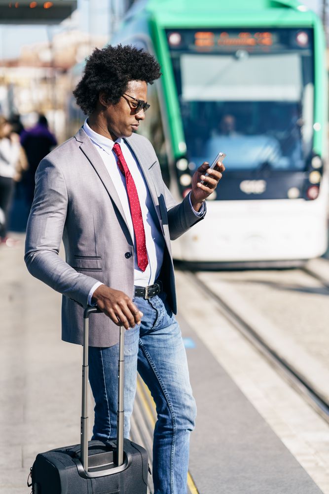 Man in jeans and dress shirt with tie standing on train platform with small suitcase