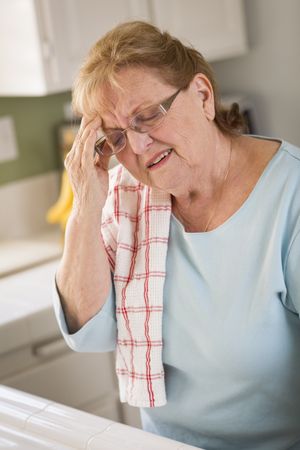 Older Adult Woman At Kitchen Sink With Head Ache
