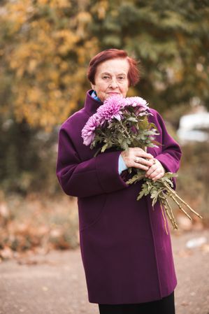 Older woman in purple coat holding bouquet of purple flowers standing near yellow trees