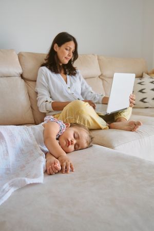 Mother using netbook while sitting on sofa near sleeping baby