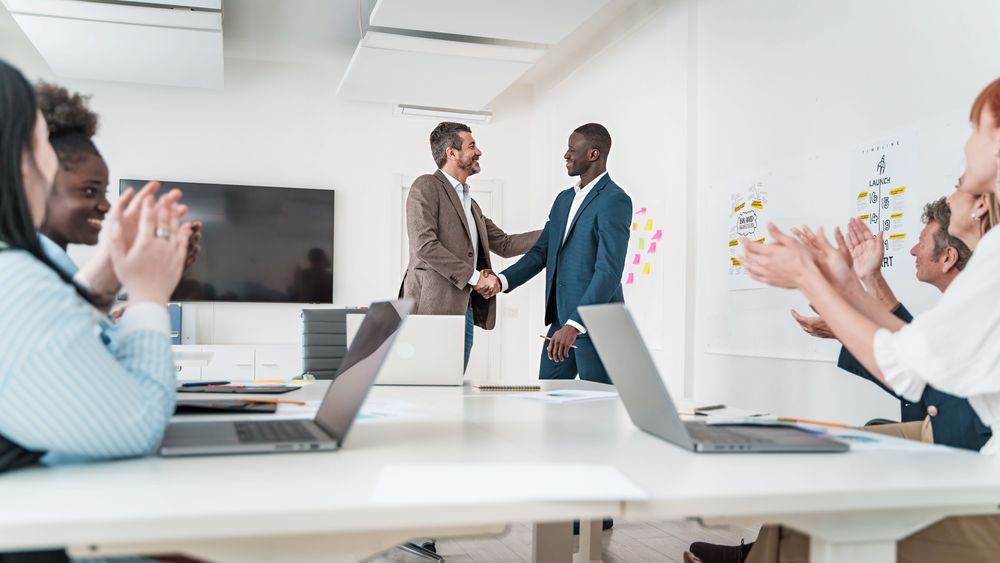 Two professionals shaking hands with colleagues clapping in the background in a bright office setting